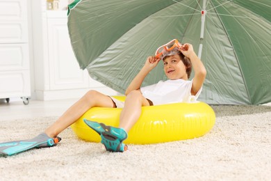 Photo of Dreaming about vacation at sea. Little boy with diving mask, flippers and inflatable ring on floor indoors