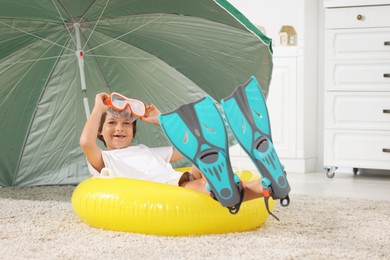Photo of Dreaming about vacation at sea. Little boy with diving mask, flippers and inflatable ring on floor indoors