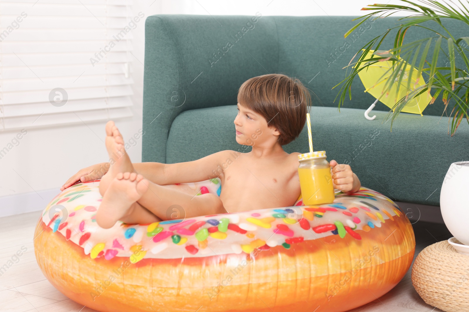 Photo of Dreaming about vacation at sea. Little boy with mason jar of fresh juice and inflatable ring on floor indoors