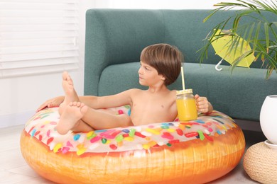 Photo of Dreaming about vacation at sea. Little boy with mason jar of fresh juice and inflatable ring on floor indoors