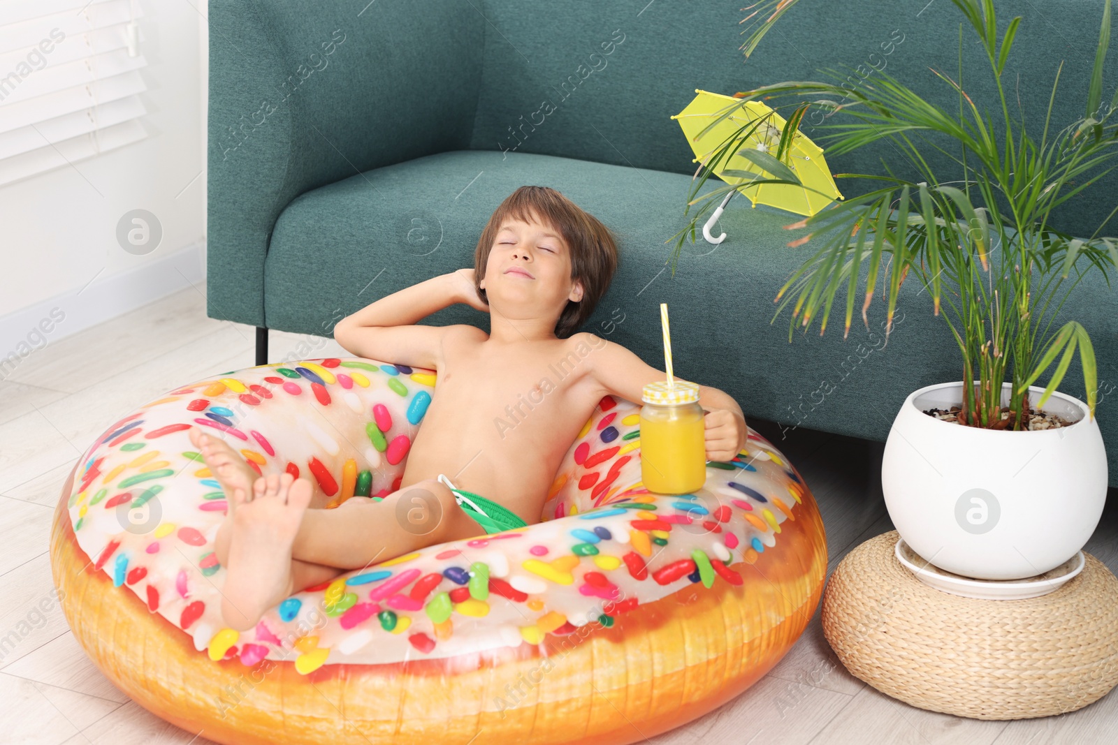 Photo of Dreaming about vacation at sea. Little boy with mason jar of fresh juice and inflatable ring on floor indoors