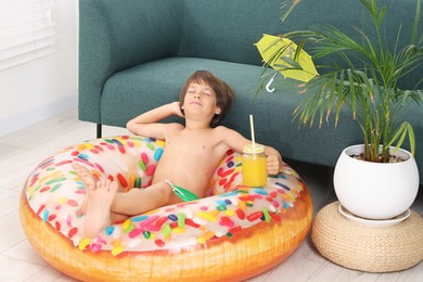 Photo of Dreaming about vacation at sea. Little boy with mason jar of fresh juice and inflatable ring on floor indoors