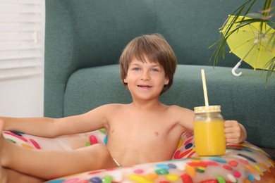 Photo of Dreaming about vacation at sea. Little boy with mason jar of fresh juice and inflatable ring on floor indoors