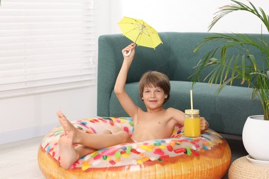 Photo of Dreaming about vacation at sea. Little boy with mason jar of fresh juice, small umbrella and inflatable ring on floor indoors