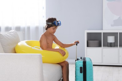 Photo of Dreaming about vacation at sea. Little boy with diving mask, flippers, inflatable ring and suitcase indoors