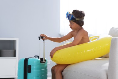 Photo of Dreaming about vacation at sea. Little boy with diving mask, flippers, inflatable ring and suitcase indoors