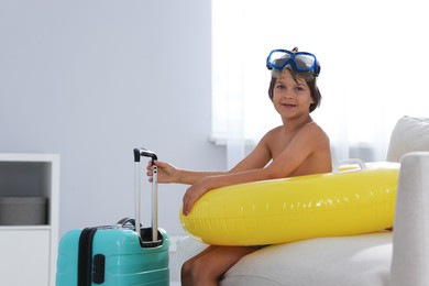 Photo of Dreaming about vacation at sea. Little boy with diving mask, flippers, inflatable ring and suitcase indoors
