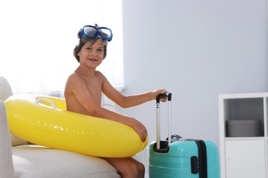 Photo of Dreaming about vacation at sea. Little boy with diving mask, flippers, inflatable ring and suitcase indoors