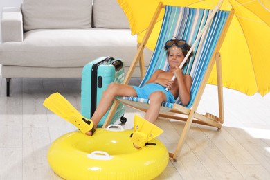 Photo of Dreaming about vacation at sea. Little boy with diving mask and flippers on deckchair near inflatable ring indoors