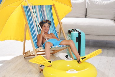 Photo of Dreaming about vacation at sea. Little boy with diving mask and flippers on deckchair near inflatable ring indoors