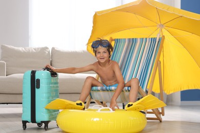 Photo of Dreaming about vacation at sea. Little boy with diving mask and flippers on deckchair near inflatable ring indoors