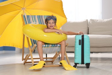 Photo of Dreaming about vacation at sea. Little boy with diving mask, flippers and inflatable ring on deckchair indoors
