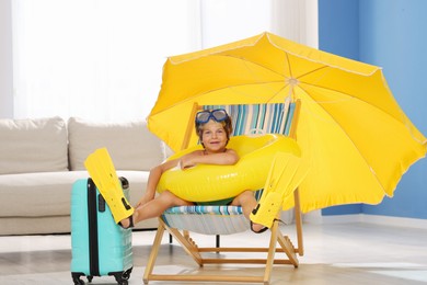 Photo of Dreaming about vacation at sea. Little boy with diving mask, flippers and inflatable ring on deckchair indoors
