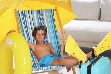 Photo of Dreaming about vacation at sea. Little boy with diving mask and flippers on deckchair near inflatable ring indoors