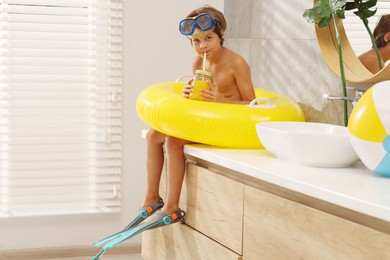 Photo of Dreaming about vacation at sea. Little boy with diving mask, juice, flippers and inflatable ring in bathroom, space for text