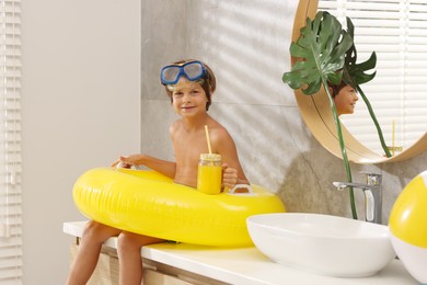 Photo of Dreaming about vacation at sea. Little boy with diving mask, juice, flippers and inflatable ring in bathroom