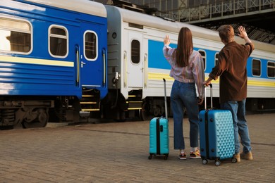 Photo of Goodbye. Couple with suitcases waving on platform of railway station, back view. Space for text
