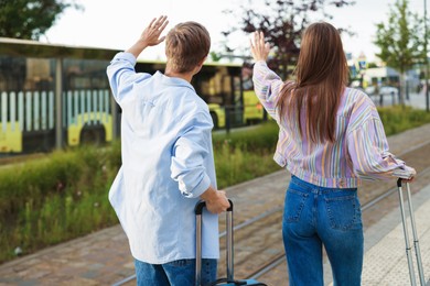 Photo of Goodbye. Couple with suitcases waving at bus stop, back view