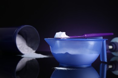 Photo of Blonde hair care cosmetic product. Powder in bowl, jar and spatula on black mirror surface against dark purple background, closeup