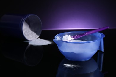 Photo of Blonde hair care cosmetic product. Powder in bowl, jar and spatula on black mirror surface against dark purple background, closeup