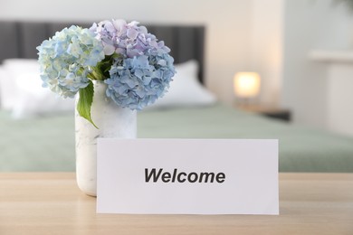 Photo of Card with word Welcome and hydrangea flowers on wooden table in hotel room