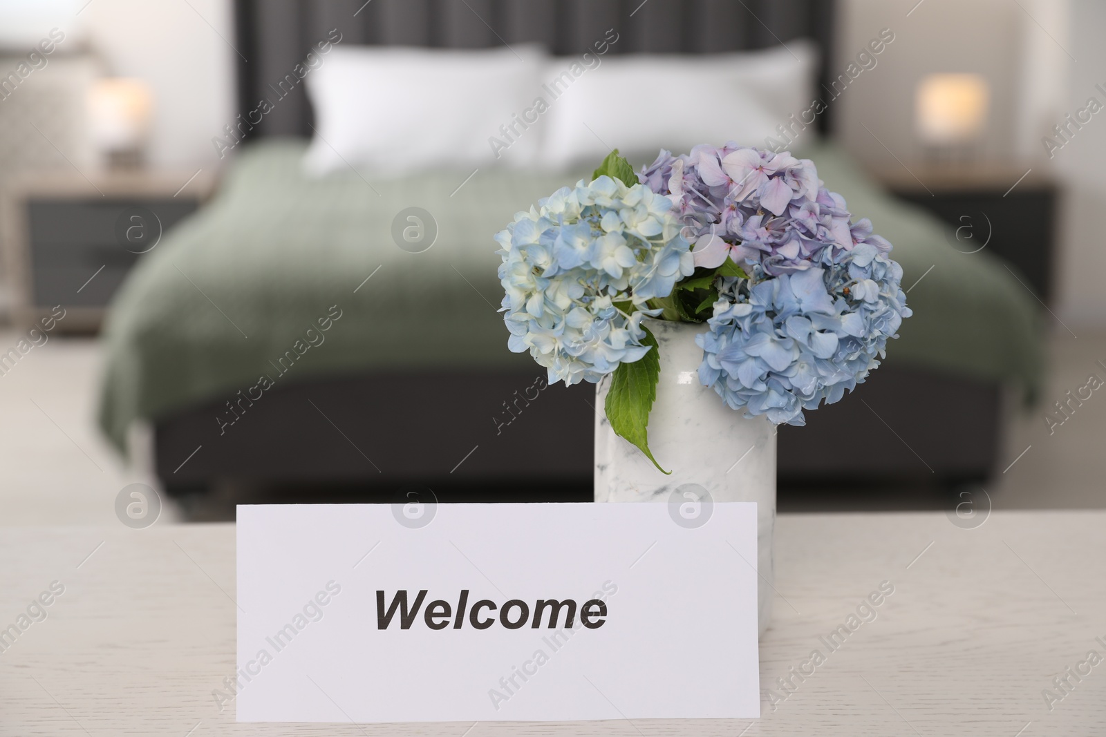 Photo of Card with word Welcome and hydrangea flowers on white wooden table in hotel room