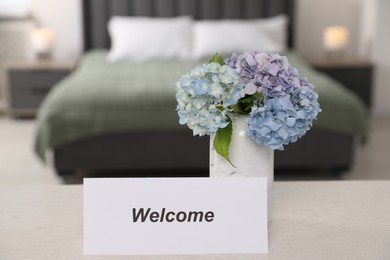 Photo of Card with word Welcome and hydrangea flowers on white wooden table in hotel room