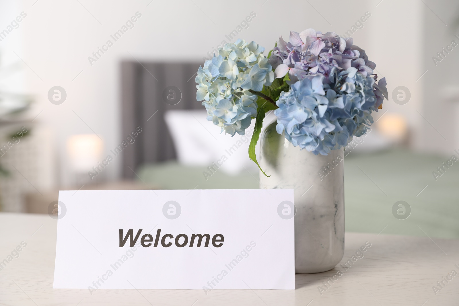 Photo of Card with word Welcome and hydrangea flowers on white wooden table in hotel room