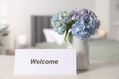Photo of Card with word Welcome and hydrangea flowers on white wooden table in hotel room
