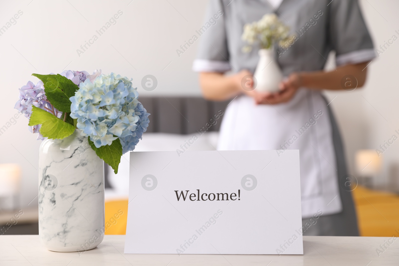 Chambermaid in hotel room, focus on card with word Welcome and hydrangea flowers on white wooden table Photo of Chambermaid in hotel room, focus on card with word Welcome and hydrangea flowers on white wooden table
