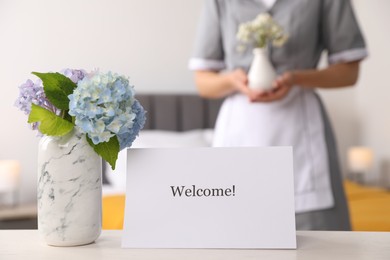 Photo of Chambermaid in hotel room, focus on card with word Welcome and hydrangea flowers on white wooden table