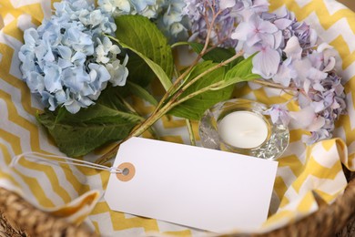 Photo of Empty card, candle and hydrangea flowers in wicker basket, closeup