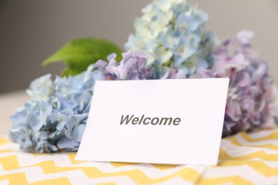Photo of Card with word Welcome and hydrangea flowers on table, closeup