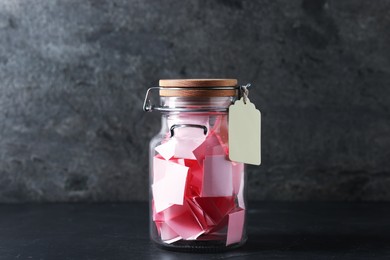 Photo of Pink paper pieces in glass jar on black table. Notes with messages