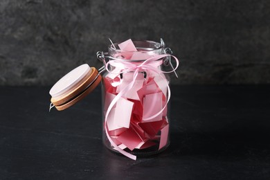 Photo of Pink paper pieces in glass jar on black table. Notes with messages