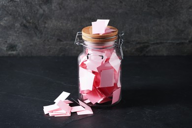 Photo of Pink paper pieces in glass jar on black table. Notes with messages