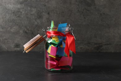 Photo of Colorful paper pieces in glass jar on black table. Notes with messages