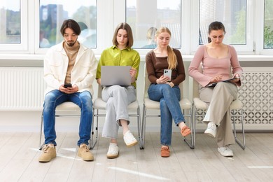 Photo of People with different devices waiting in queue on chairs indoors