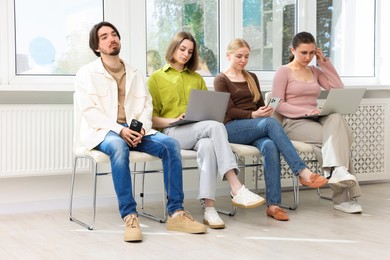 Photo of People with different devices waiting in queue on chairs indoors