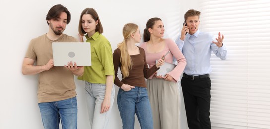Photo of People with different devices waiting in queue near white wall indoors