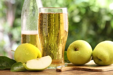 Photo of Delicious cider in glass, bottle, apples and green leaves on wooden table outdoors, closeup