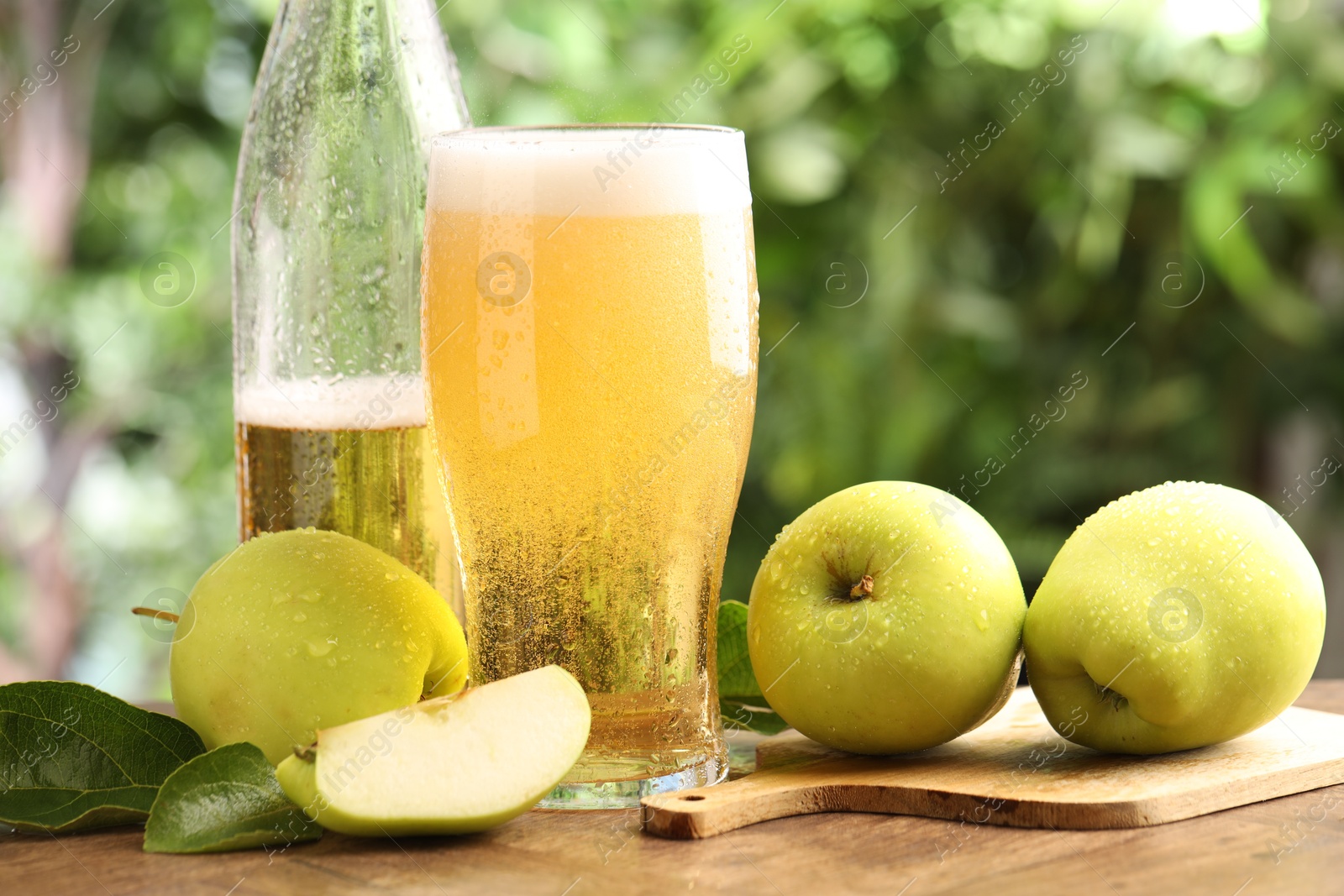 Photo of Delicious cider in glass, bottle, apples and green leaves on wooden table outdoors