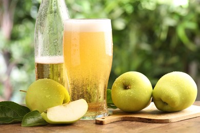 Photo of Delicious cider in glass, bottle, apples and green leaves on wooden table outdoors