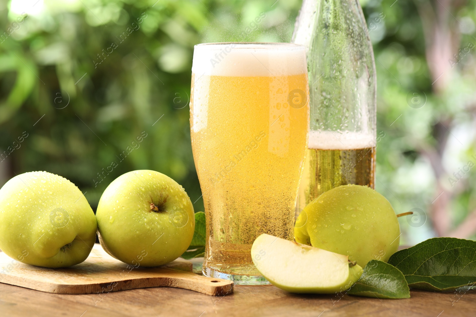 Photo of Delicious cider in glass, bottle, apples and green leaves on wooden table outdoors