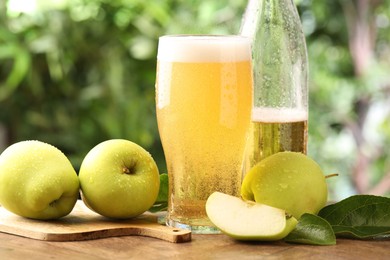Photo of Delicious cider in glass, bottle, apples and green leaves on wooden table outdoors