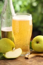 Photo of Delicious cider in glass, bottle, apples and green leaves on wooden table outdoors, closeup