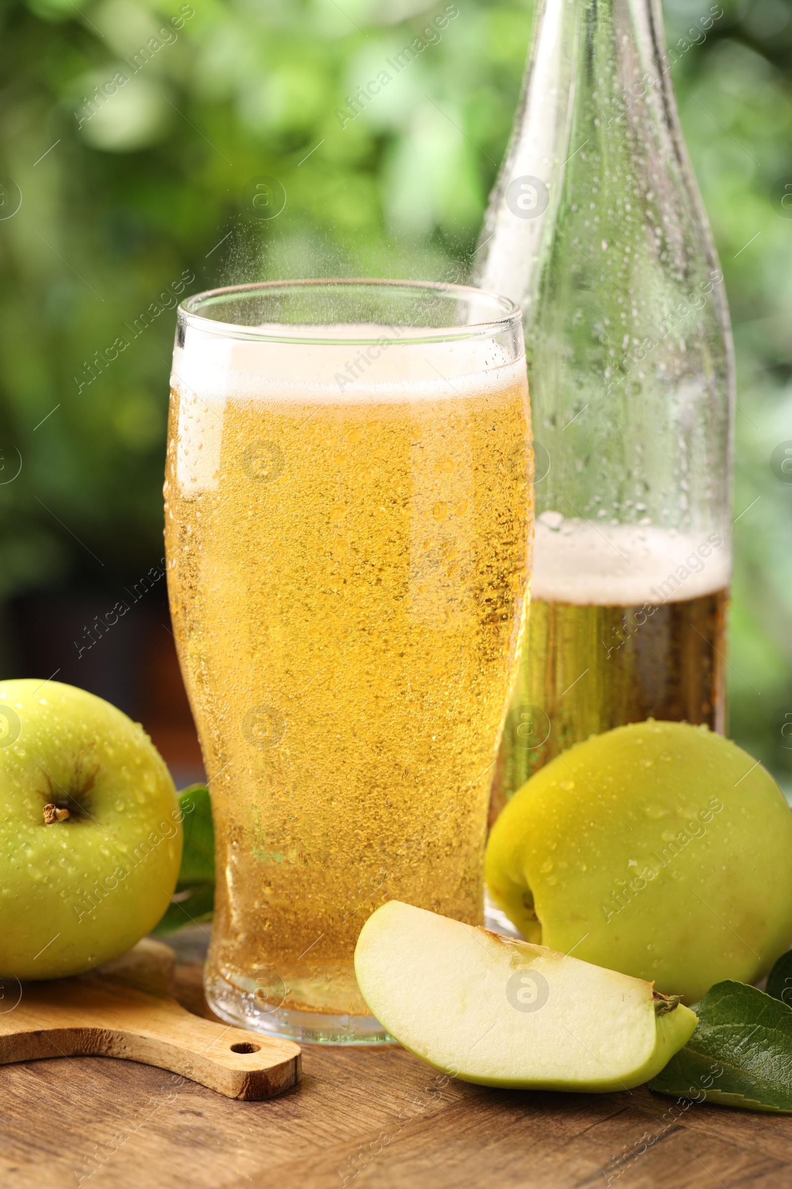 Photo of Delicious cider in glass, bottle, apples and green leaves on wooden table outdoors, closeup