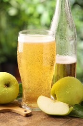 Photo of Delicious cider in glass, bottle, apples and green leaves on wooden table outdoors, closeup