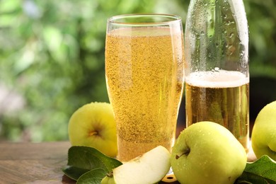 Photo of Delicious cider in glass, bottle, apples and green leaves on wooden table outdoors, closeup. Space for text