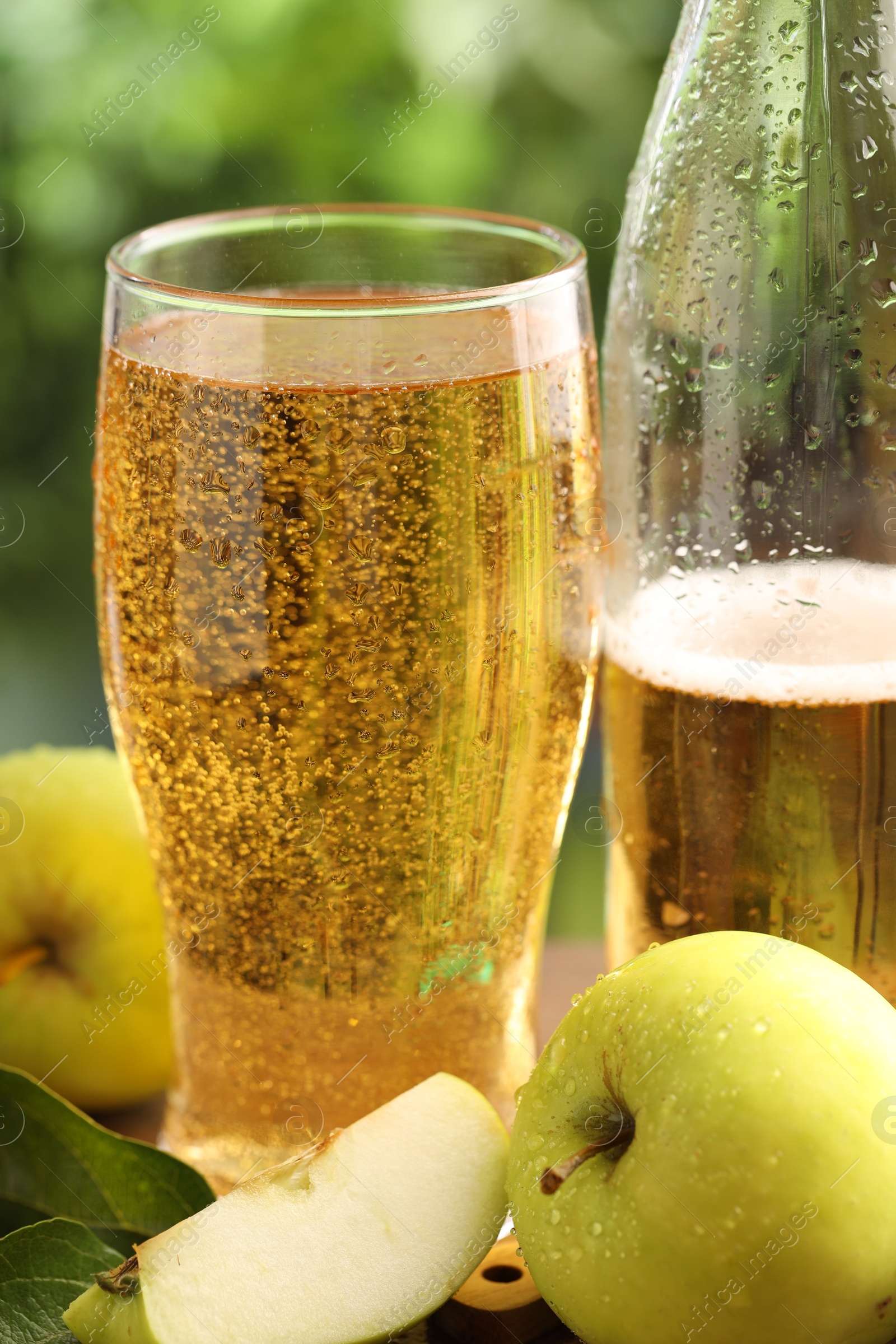 Photo of Delicious cider in glass, bottle, apples and green leaves on table outdoors, closeup